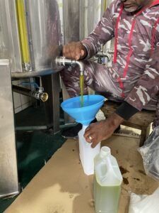Worker in camo jumpsuit opens a tap on a stainless steel tank, pouring green liquid into a blue funnel over a white jugBelow.