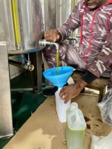 Worker pours green liquid from a stainless steel tank through a blue funnel into a white container.